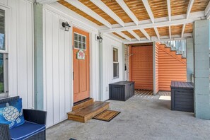 Eine überdachte Veranda mit Holzbalkendecke, eine Tür mit rotem Rahmen und eine Treppe, die zu einer Garage führt.