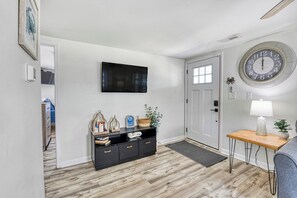 A hallway with a wooden floor, a black console table with decorative items, a round wall clock, and a door with a window. - Cheeky Crab B - 2 Bedroom Coastal Cottage Steps from the Beach (Garden City Beach)