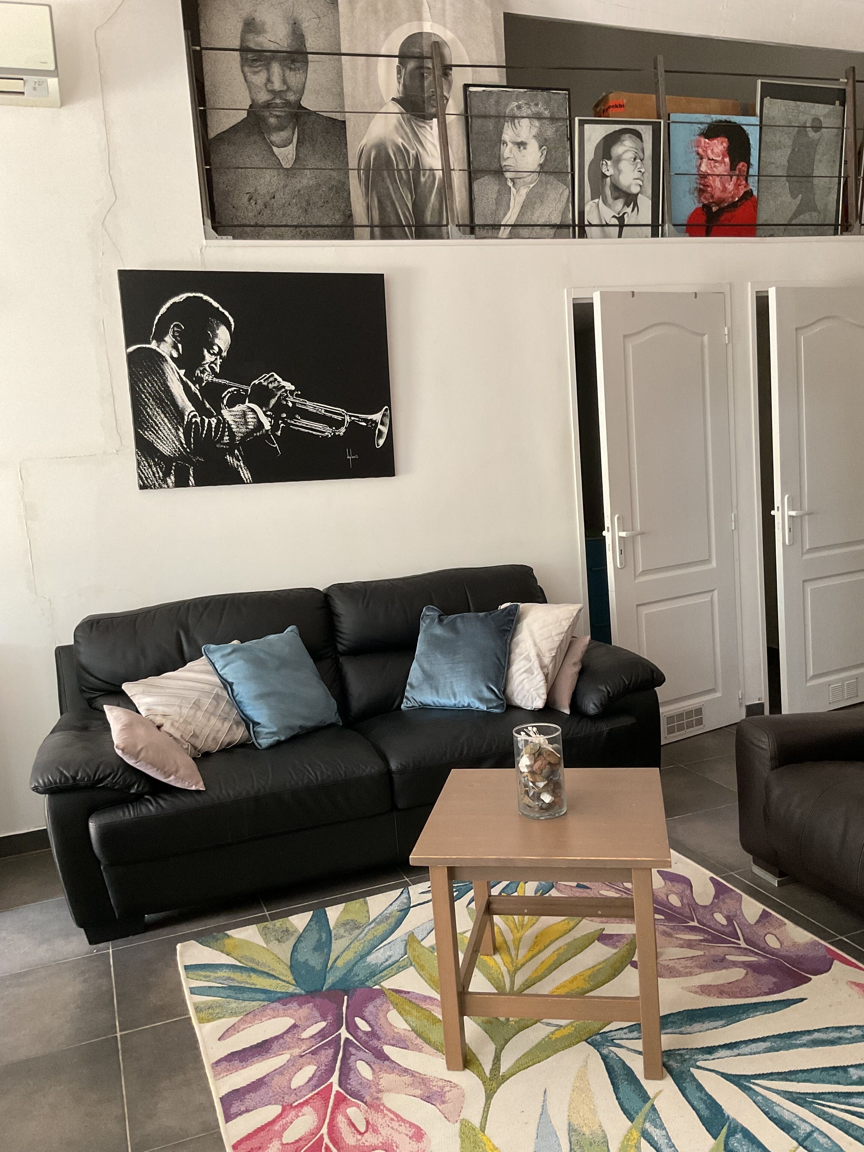 A living room with a black leather sofa, a coffee table, and a colorful rug. There are framed pictures on the wall and a door in the background.