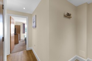 A hallway with a wooden floor, beige walls, a door leading to a room, and a small shelf with decorative items. - Heathrow Living 2BDR 2BA Apt.12 (Stanwell)
