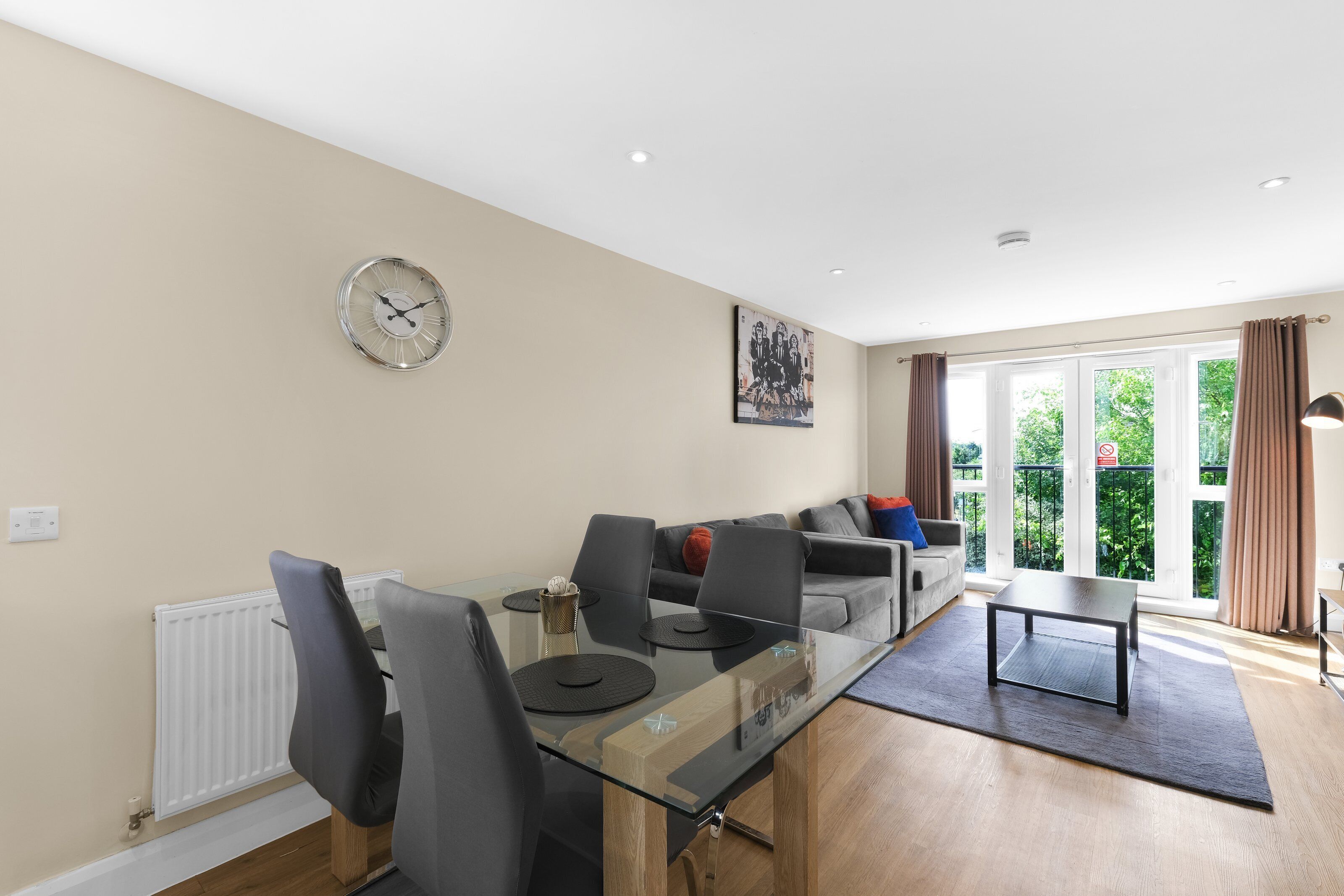 A modern living room with a glass dining table, grey sofas, and a large window with curtains.