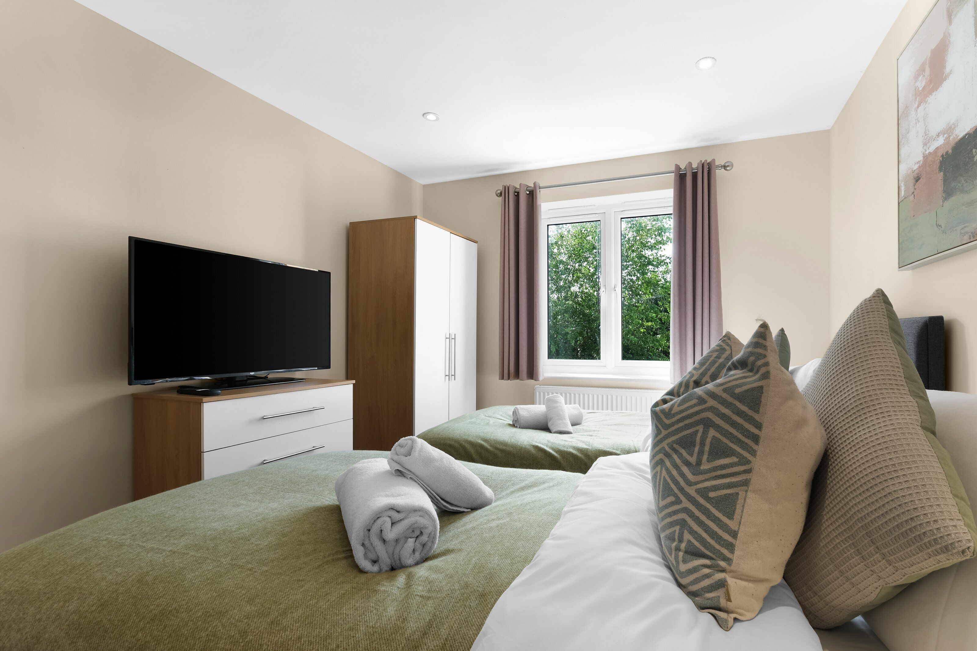 A hotel room with a flat-screen TV, two beds with green bedding, a wooden wardrobe, and a window with curtains.