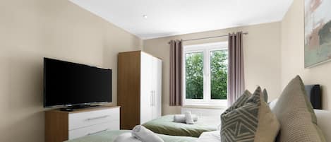 A hotel room with a flat-screen TV, two beds with green bedding, a wooden wardrobe, and a window with curtains.