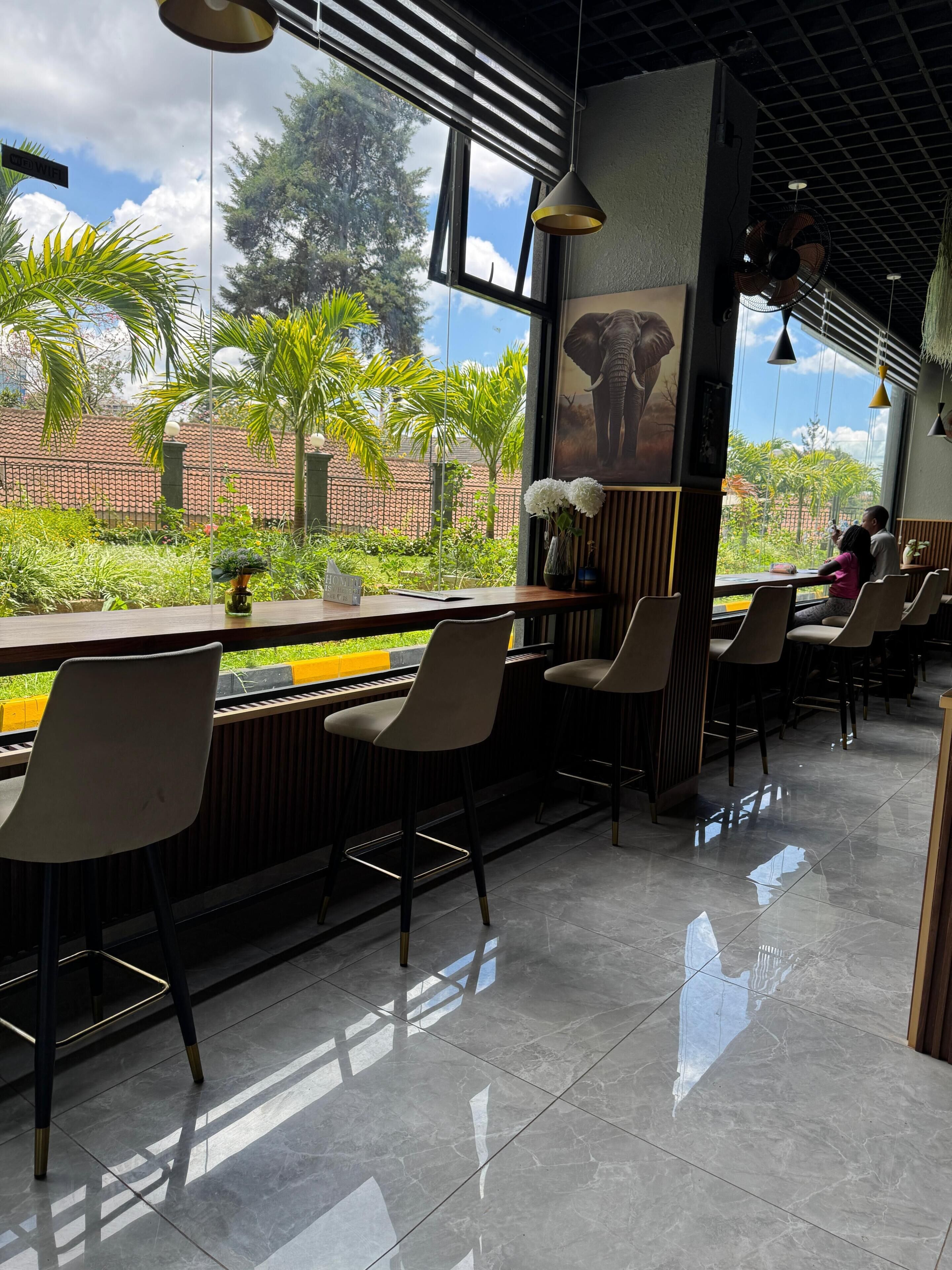 A restaurant with a view of a garden, featuring a long counter with chairs, a tiled floor, and a large window.