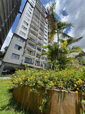 A multi-story residential building with balconies, surrounded by a garden with yellow flowers.
