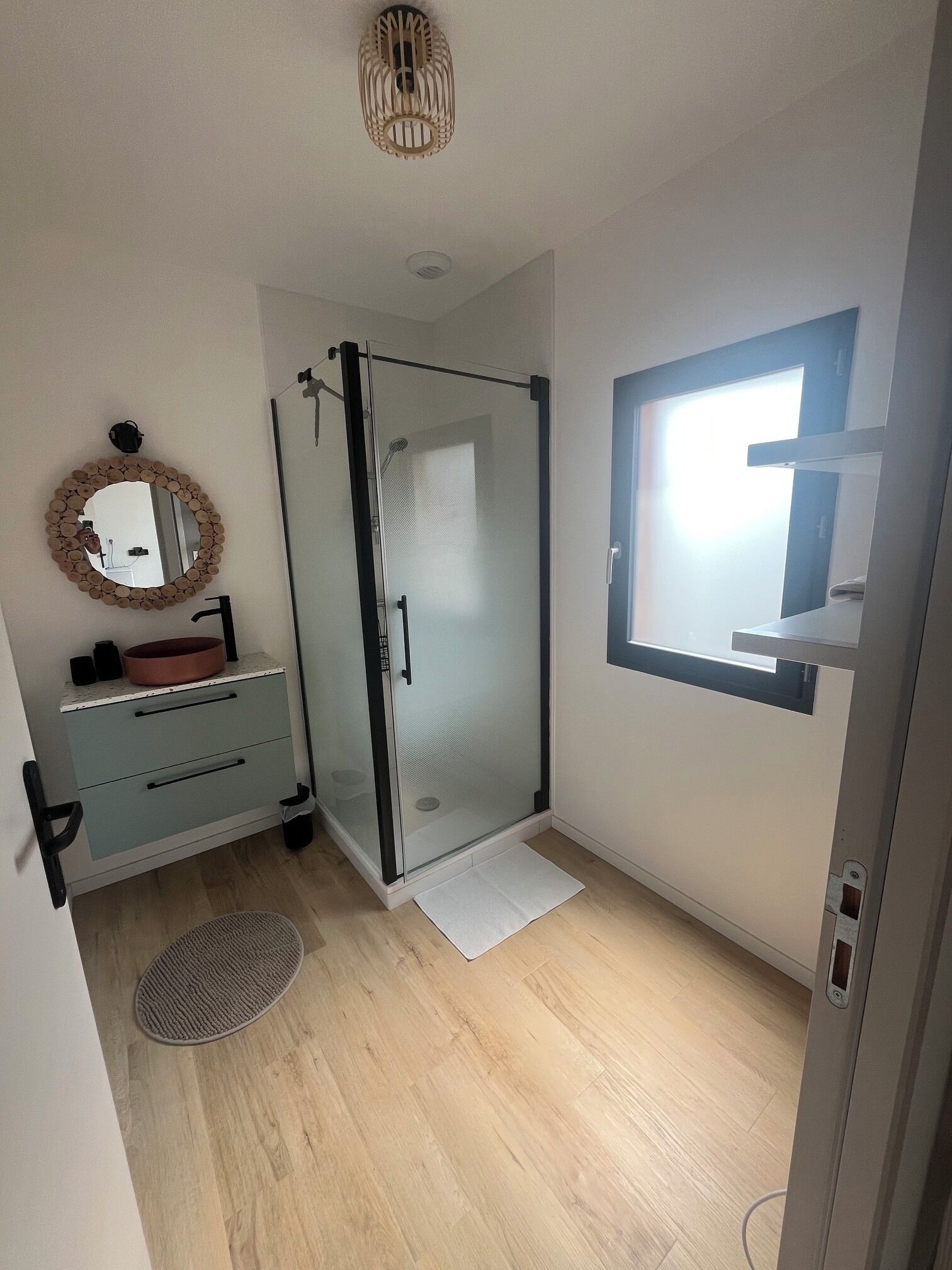 Un baño moderno con cabina de ducha de vidrio, un espejo redondo con marco de madera y un lavamanos pequeño con lavabo de cobre.