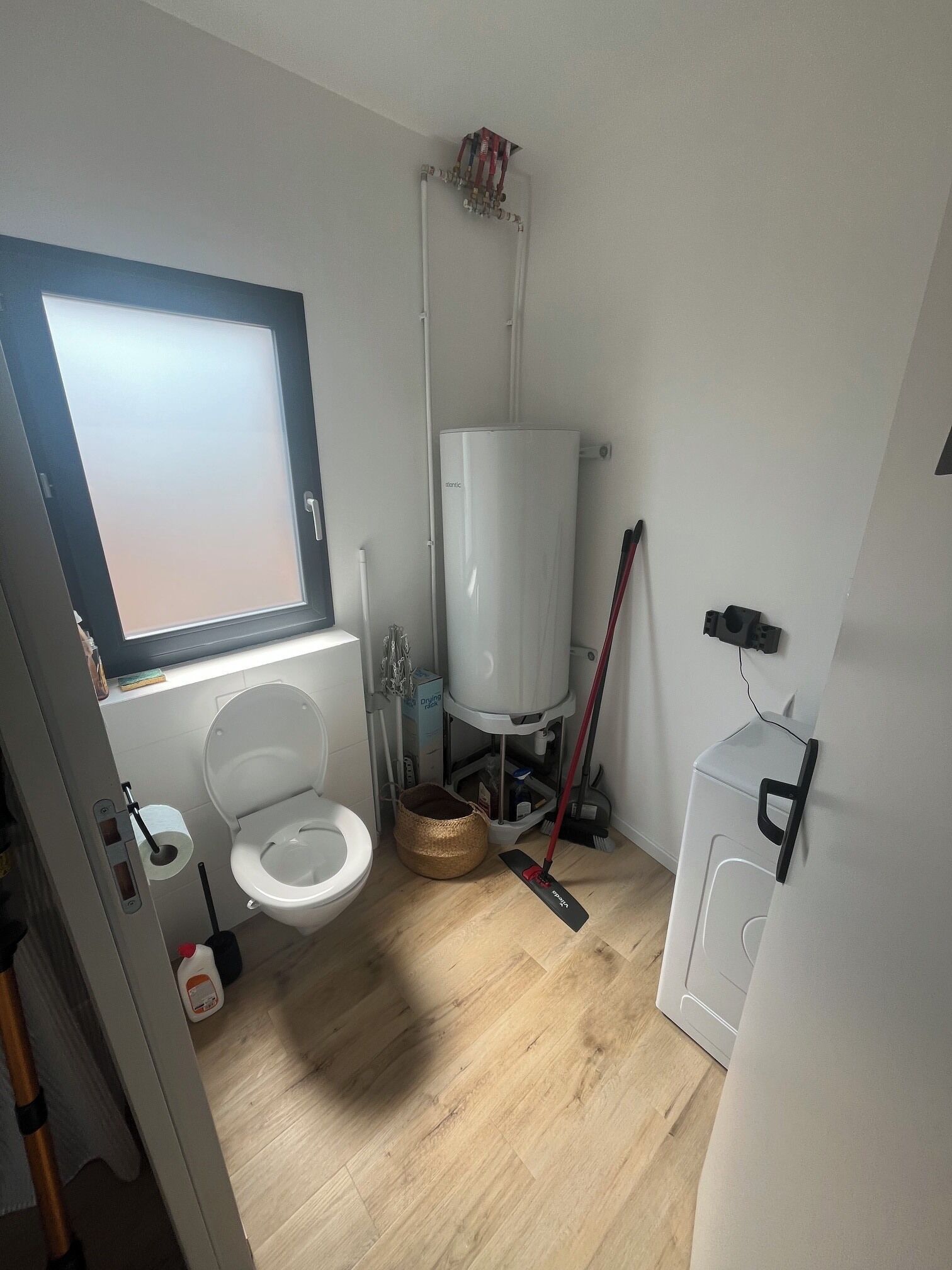 A small, compact bathroom with a toilet, a window, a water heater, and a washing machine.