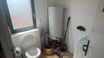 A small, compact bathroom with a toilet, a window, a water heater, and a washing machine.