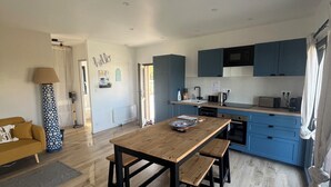 A modern kitchen with blue cabinets, a wooden dining table with benches, and a visible window with curtains. - BELLEVUE COTTAGE (Tréveneuc)