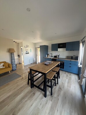 A modern kitchen with blue cabinets, a wooden dining table with benches, and a visible window with curtains. - BELLEVUE COTTAGE (Tréveneuc)