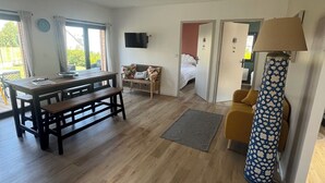 A spacious room with a wooden floor, a dining table with benches, a sofa, a TV mounted on the wall, and a lamp. - BELLEVUE COTTAGE (Tréveneuc)
