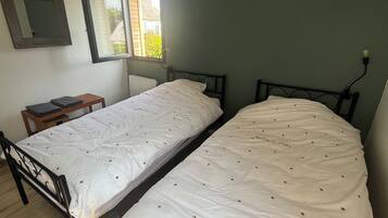 Ein Zimmer mit zwei Einzelbetten, einem kleinen Tisch, einer Hängeleuchte und einem Fenster mit Blick auf ein Haus.