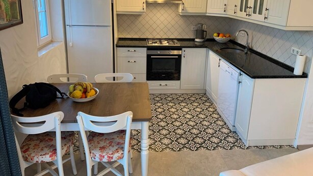 Een compacte keuken met witte kastjes, een Siemens koelkast, een fornuis en een spoelbak. Er staat een tafel met stoelen en een schaal met fruit.