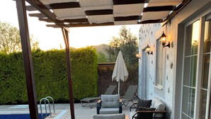Een terras buiten met een eettafel voor twee personen, omringd door rieten stoelen, een zwembad en een overdekt zithoekje met banken en een tafel.