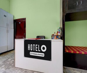 Ein Hotelrezeptionstresen mit einem Schild, das darauf hinweist, dass er vom Unternehmen gebucht wurde. Im Hintergrund ist eine rote Tür und ein Bett mit einem gemusterten Überwurf zu sehen.