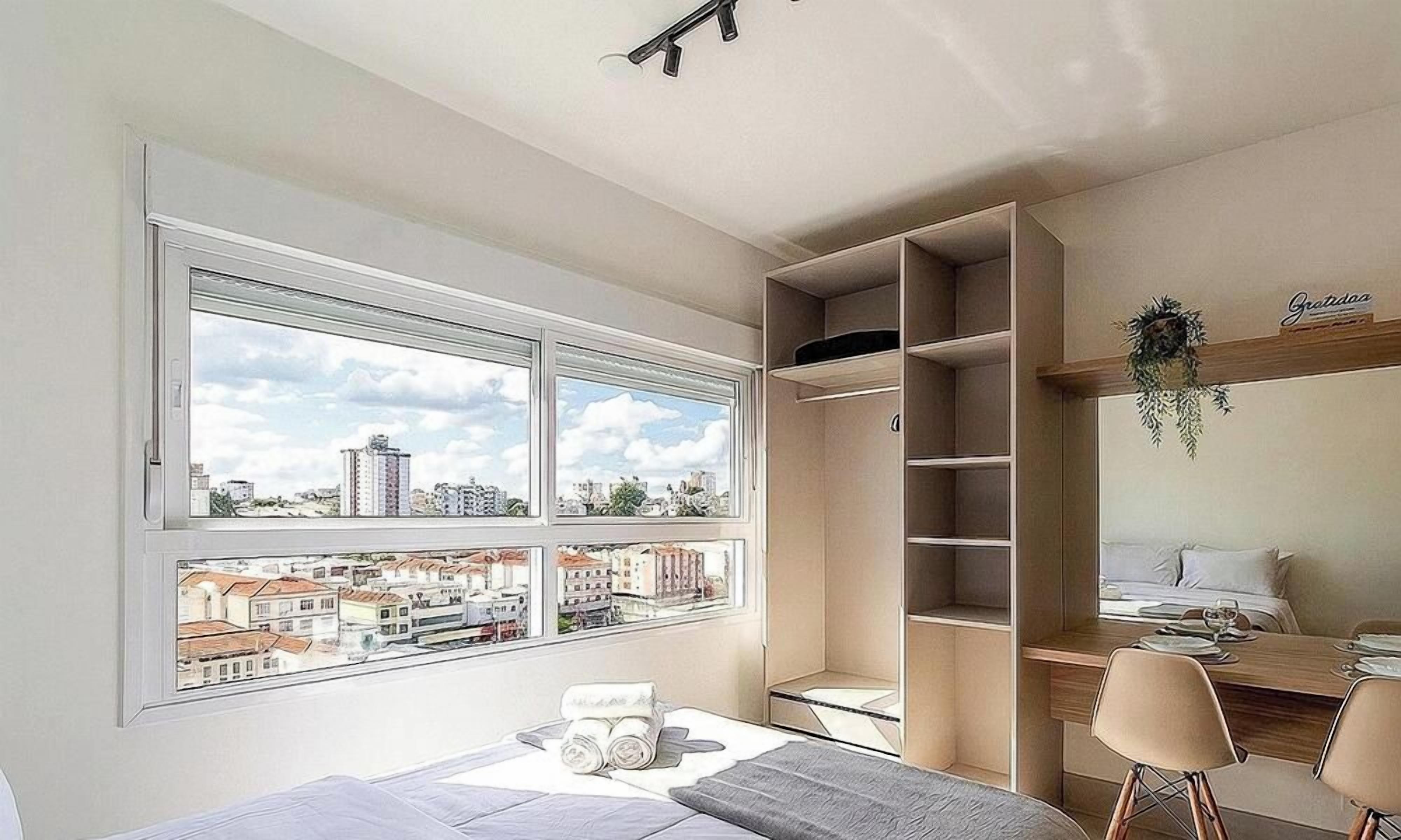 Quarto com uma janela grande, cama, escrivaninha com cadeiras e estantes embutidas.