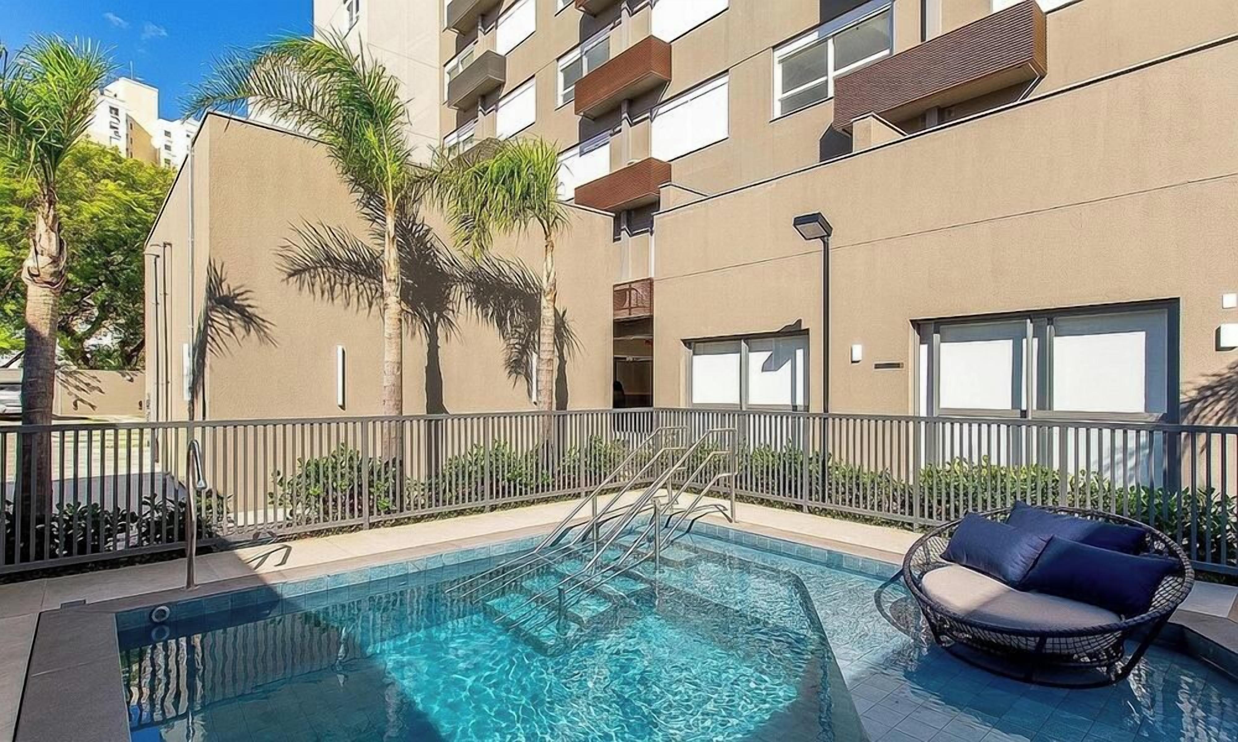 Piscina com escada de metal e cadeira de vime, cercada por uma grade e palmeiras.