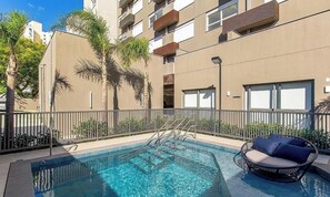 Ein Swimmingpool mit Metallleiter und einem Korbstuhl, umgeben von einem Zaun und Palmen.