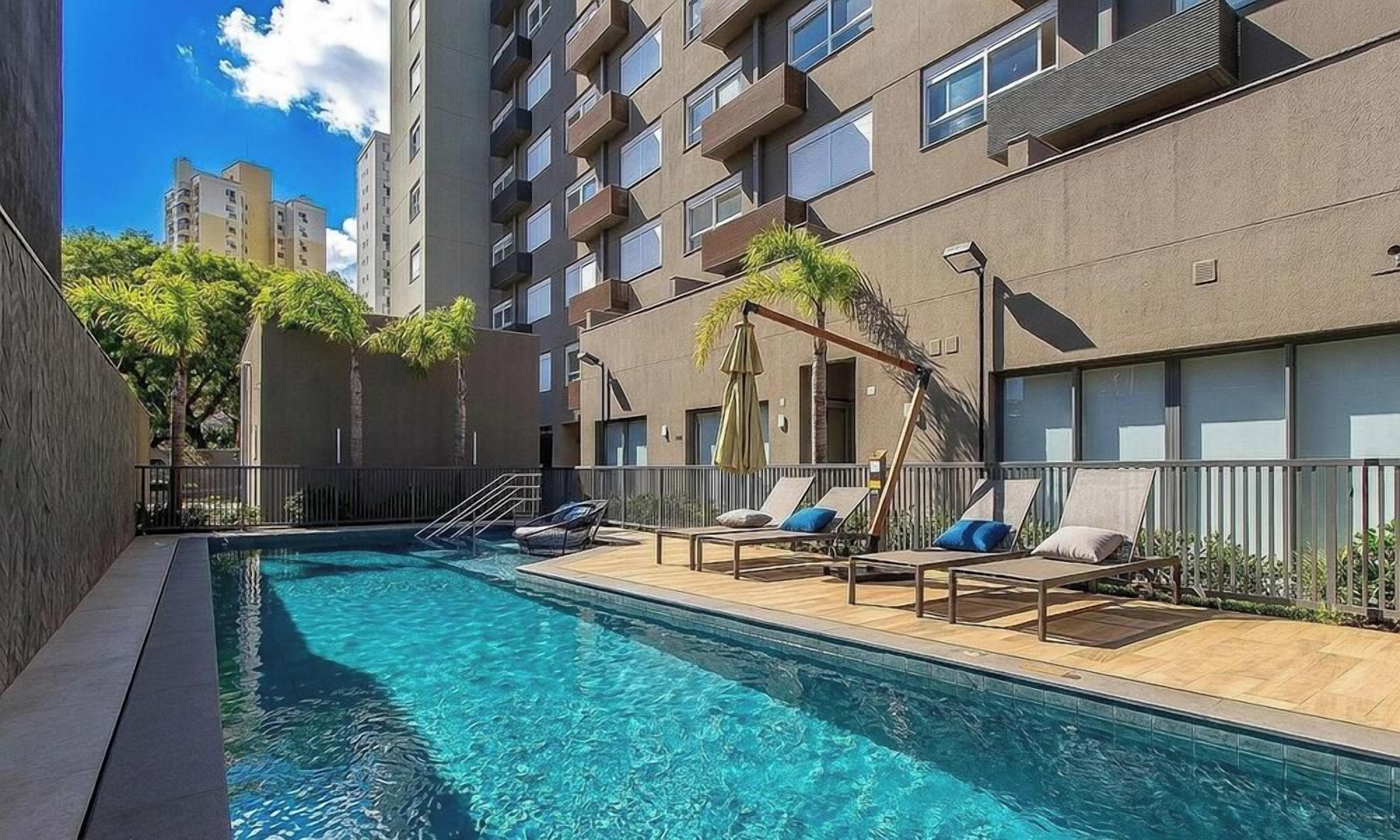 Uma piscina com espreguiçadeiras e guarda-sóis, cercada por um edifício com varandas e janelas.