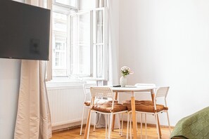 Ein Zimmer mit einem weißen Tisch und Stühlen, einer Blumenvase und einem großen Fenster mit weißen Fensterläden.