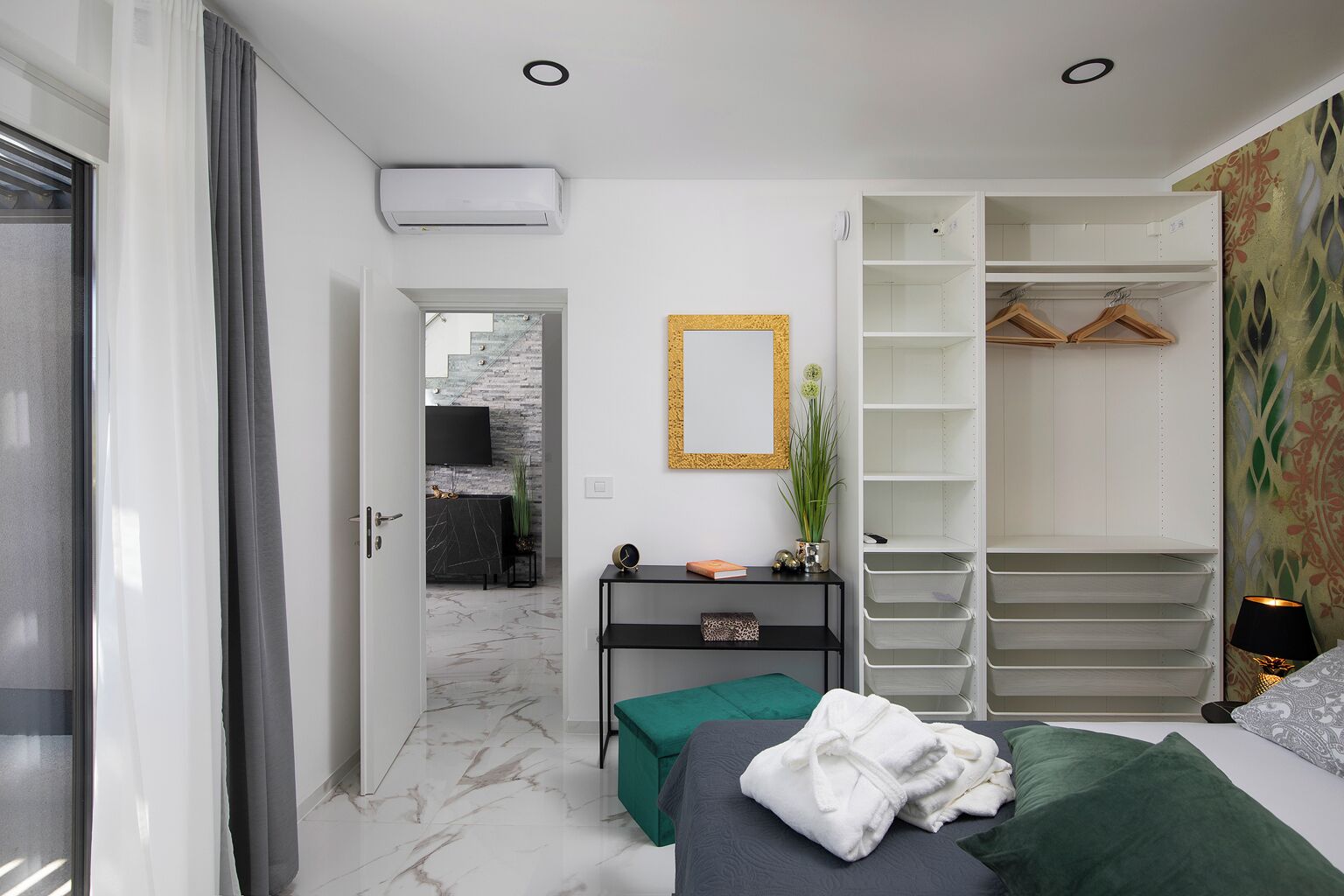 Une chambre à coucher comprenant un lit, une table de chevet, un miroir, une armoire et une salle de bain visible à travers une porte ouverte.