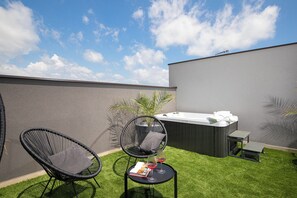 Eine Dachterrasse mit zwei schwarzen Korbstühlen, einem kleinen runden Tisch mit einer Zeitschrift und zwei Weingläsern, einem Whirlpool und einer Bank.