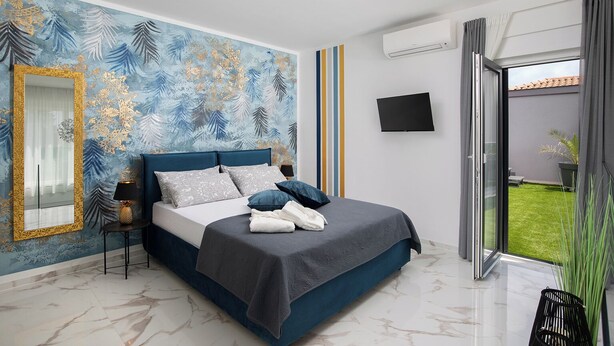 A modern bedroom with a large bed, a mirror, a flat-screen TV, and a view of a green lawn. - Villa PalmDora – New Vacation Home with Private Pool & Hot Tub in Istria (Marčana)