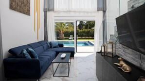A modern living room with a dark blue sofa, a glass coffee table, and a large flat-screen TV mounted on a stone-textured wall. - Villa PalmDora – New Vacation Home with Private Pool & Hot Tub in Istria (Marčana)