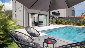 A modern outdoor pool area with a glass door entrance, lounge chairs, and a table with glasses. - Villa PalmDora – New Vacation Home with Private Pool & Hot Tub in Istria (Marčana)