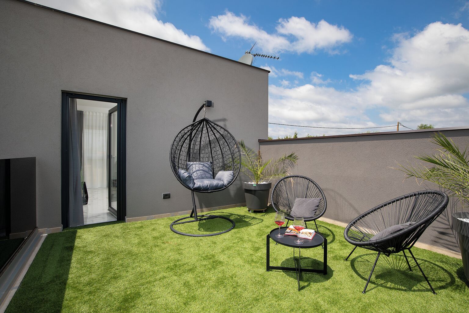 Un área de estar moderna al aire libre con dos sillones de mimbre negro, una mecedora colgante y una mesita con un vaso y un libro.