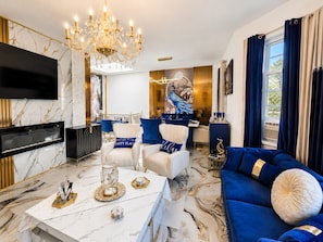 A modern living room with a marble floor, a large chandelier, a flat-screen TV, a blue sofa, and a dining area with blue chairs. - Royal 3-bedroom apartment/Wifi/15 min NYC/Bus stop 2 min/8 people (West New York)