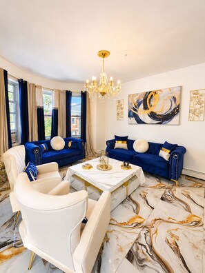 Ein luxuriöses Wohnzimmer mit einer blauen Sofagarnitur, einem weißen Sessel und einem Boden mit Marmoroptik.