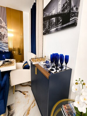 Ein modernes Wohnzimmer mit einem weißen Sofa, einem blauen Akzentstuhl, einer dunkelblauen Bar mit Gläsern und einem Stadtbild an der Wand.