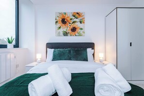 Ein modernes Hotelzimmer mit einem Bett, zwei Nachttischlampen, einer grünen Tagesdecke, einem Gemälde mit Sonnenblumen und einem Kleiderschrank.