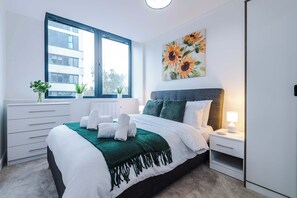 A modern hotel bedroom with a large bed, a nightstand, a window with blinds, a radiator, and a painting of sunflowers. - NEW! Chic City Base - Old Trafford & MediaCityUK (Stretford)