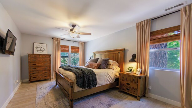 Une chambre à coucher avec un lit en bois, un ventilateur de plafond, une table de chevet, une commode et une fenêtre avec des rideaux.