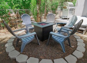 Eine Terrasse mit vier Adirondack-Stühlen, einem runden Tisch und einem Grill.