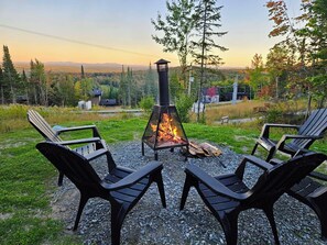 En eldstad med fyra stolar runt om, placerad på ett grusunderlag med utsikt över en skog och avlägsna berg.