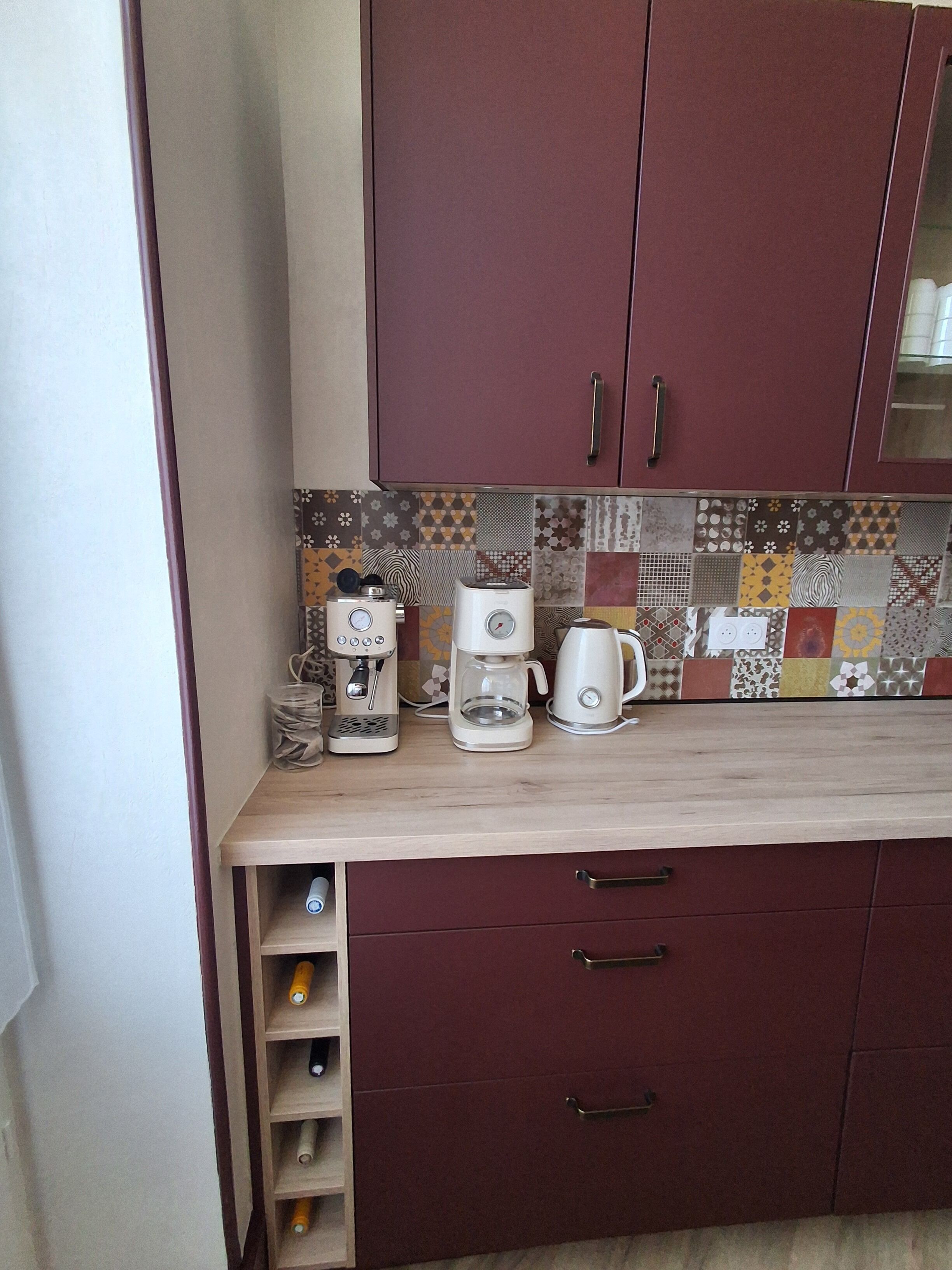 A kitchen with a coffee maker, a kettle, and a microwave on the counter. There are cabinets with handles and a tiled backsplash.