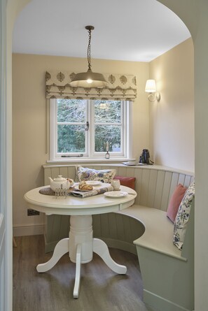 A dining area with a round table, bench seating, a window with a view of trees, and a hanging light fixture. - Ware Lodge at Woodhall Park | Sleeps 3 (Hetford)