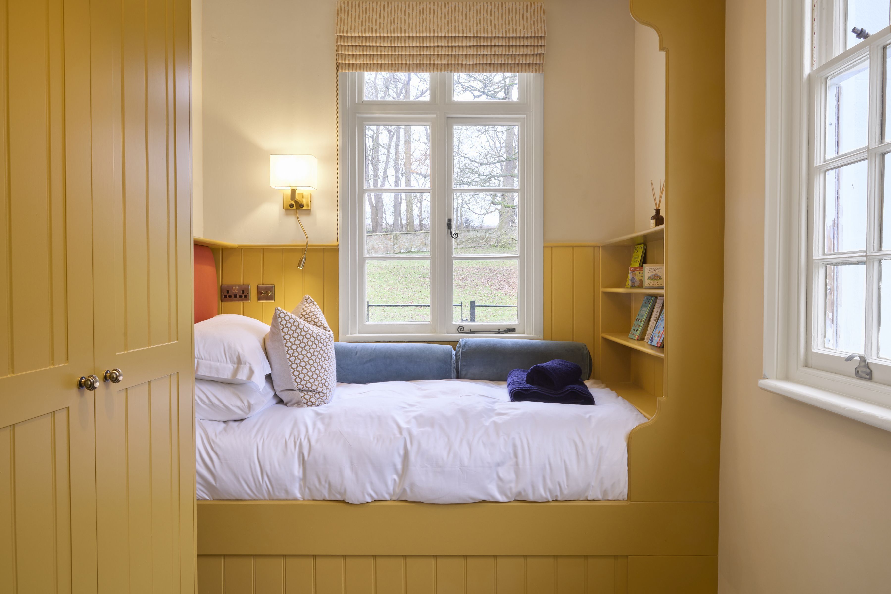 A bedroom with a bed, a window, a bedside table, and a shelf with books.