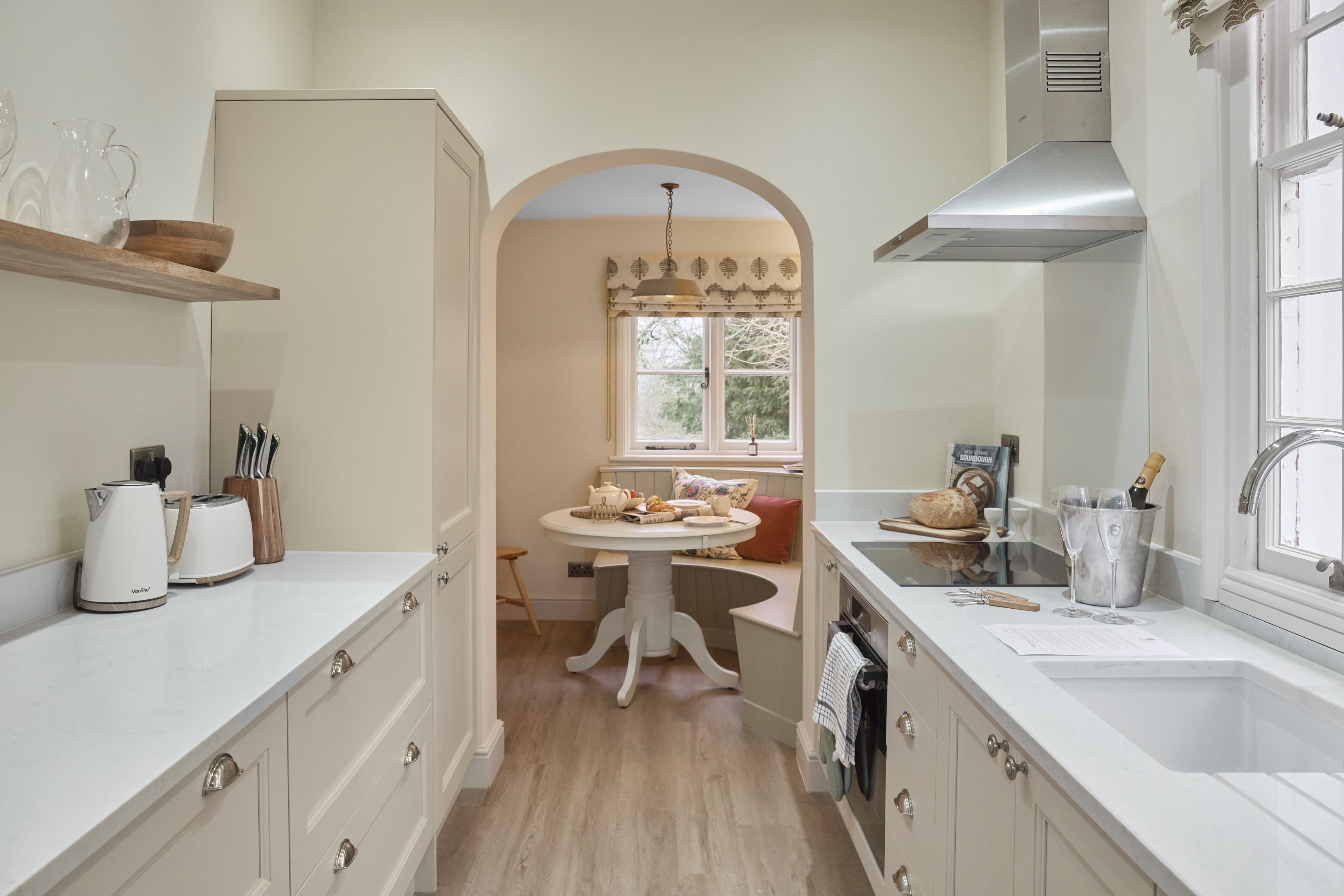A kitchen with white cabinetry, a wooden countertop, a dining table with chairs, and a window with a view of trees.
