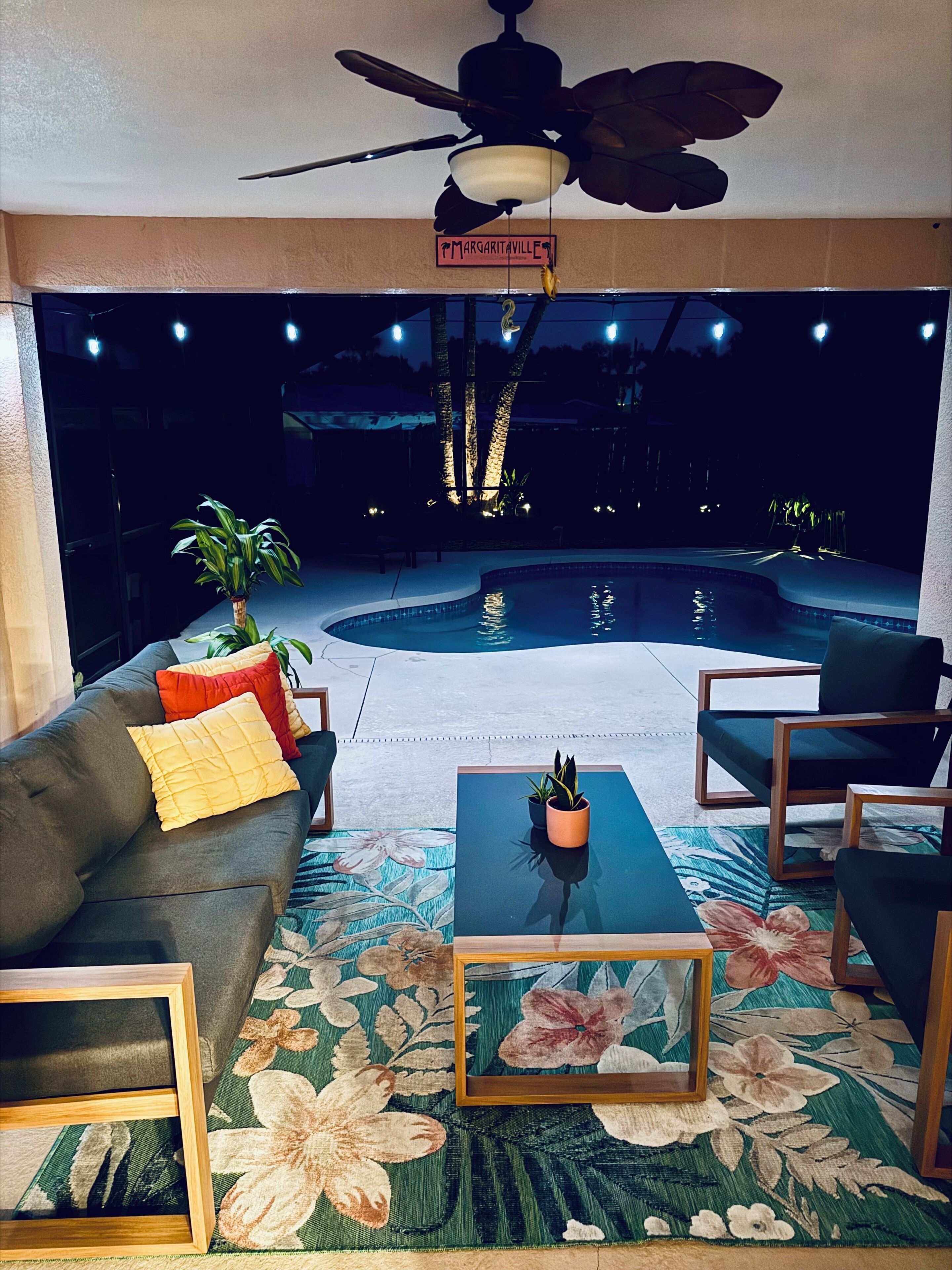 Ein Poolbereich mit Deckenventilator, einem Schild mit der Aufschrift 'Margaritaville', einer Couch mit Kissen, einem Couchtisch mit Pflanze und Sitzgelegenheiten im Freien.