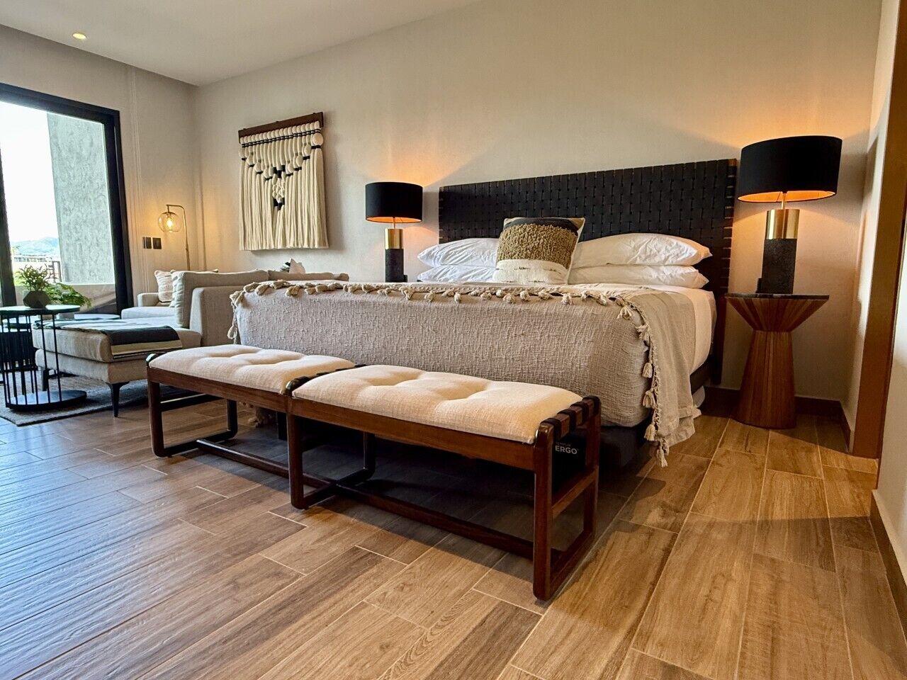 A hotel room with a bed, two bedside lamps, a bench, a sofa, and a view of the outdoors.