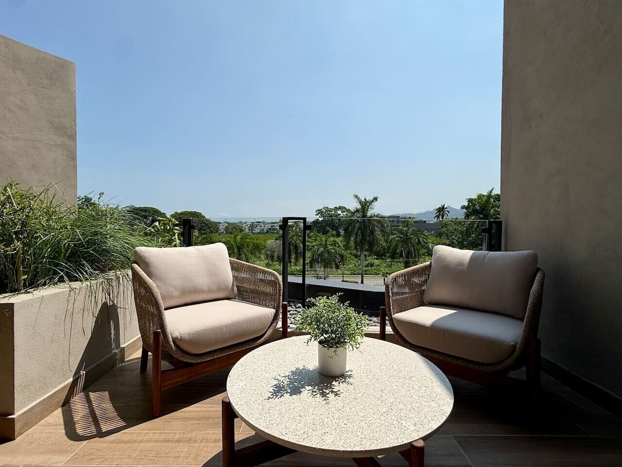 A balcony with two wicker chairs and a round table, a potted plant, and a view of greenery and buildings.