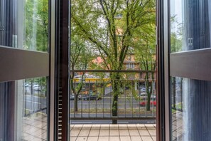 A view from a window with a metal railing, looking out onto a street with trees and parked cars. - SA7 [Island-Vertical Forest] • Large Apartment • (Milano)