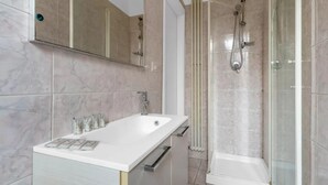Een badkamer met een wit wastafelmeubel, een spiegel, een douche met glazen deur en behangplaten in beige tinten.