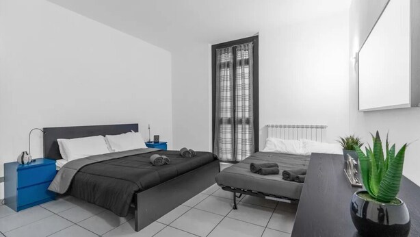 Een moderne slaapkamer met een groot bed, een kleiner bed, een bureau, een blauwe nachtkastje en een kamerplant.
