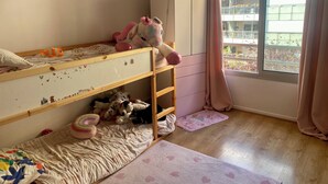 Una cama litera con juguetes, un armario blanco, una ventana con cortinas y vistas a un balcón.