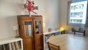 Un comedor con una mesa redonda, sillas de madera, un aparador con puertas de cristal, una lámpara y una ventana con vistas a edificios.